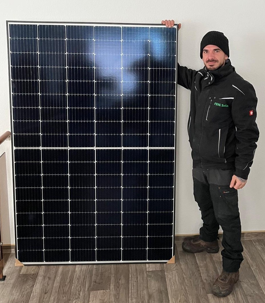 Eine Person steht lächelnd neben einem großen Solarpanel, trägt eine schwarze Jacke und Mütze.