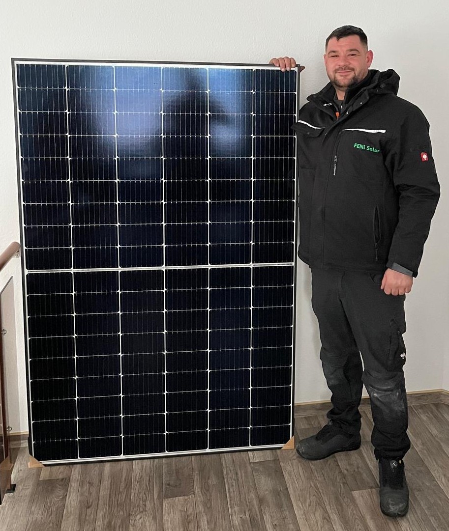 Ein Mann lächelt neben einem großen, dunklen Solarpanel in einem Raum mit Holzboden.