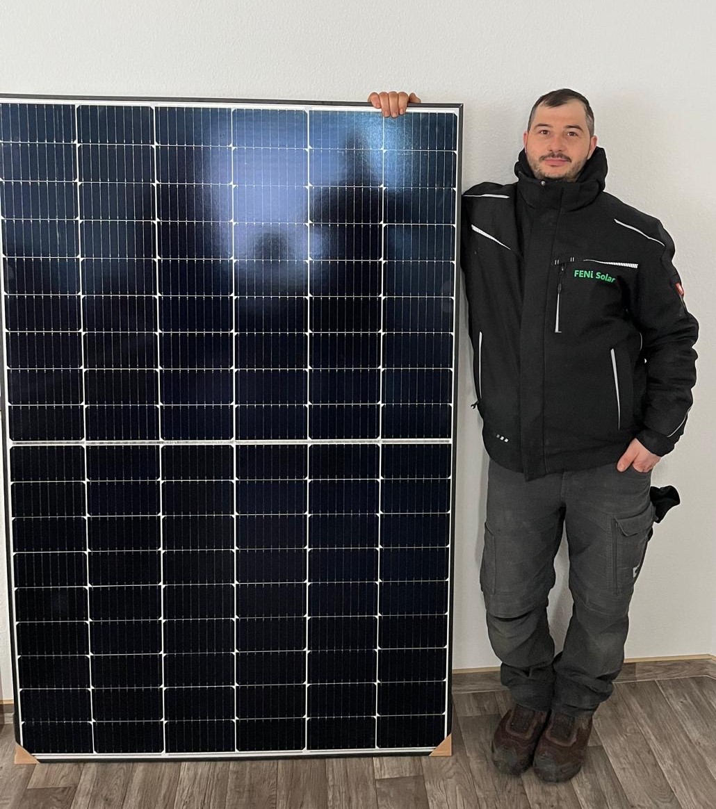 Mann steht lächelnd mit schwarzem Solar-Panel in schwarzer Jacke; Holzbodenhintergrund.